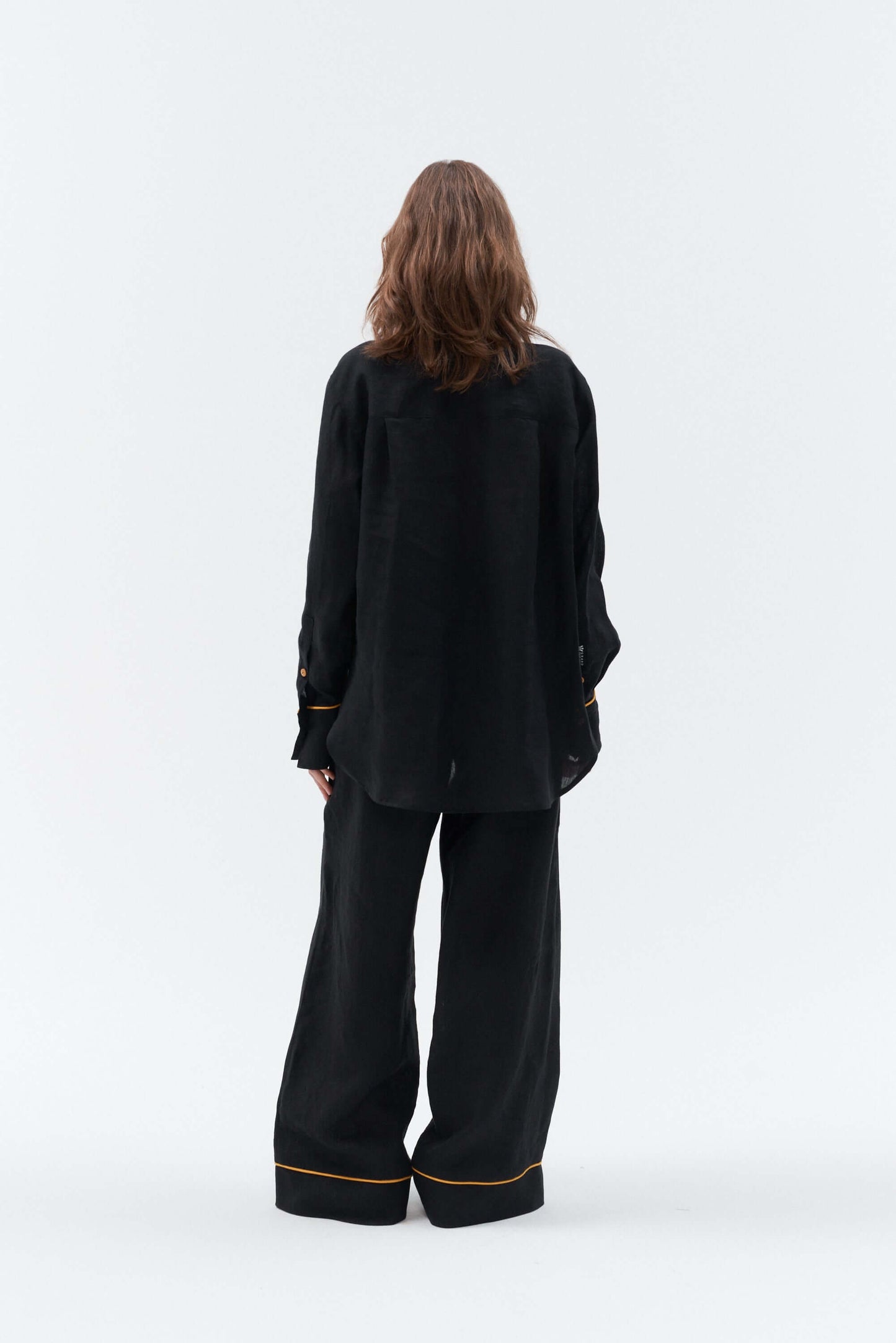 Back side of women wearing an oversized, long sleeve black linen shirt Brillante with a yellow details matching with wide-leg pants on a white background.