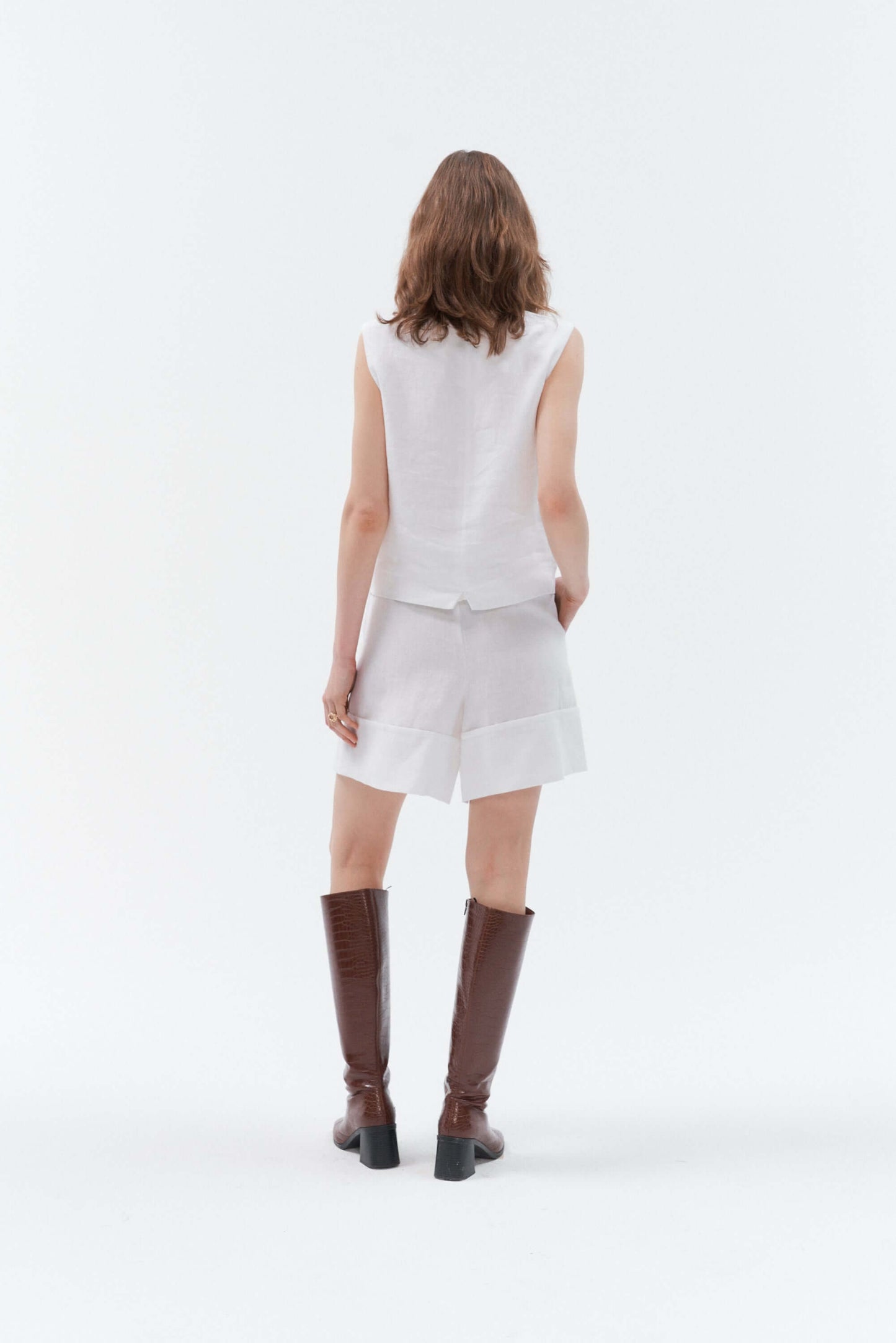 Back view of woman wearing white wide-leg linen shorts Bonita paired with the white linen waistcoat Vesta, styled with the brown high-heeled boots.
