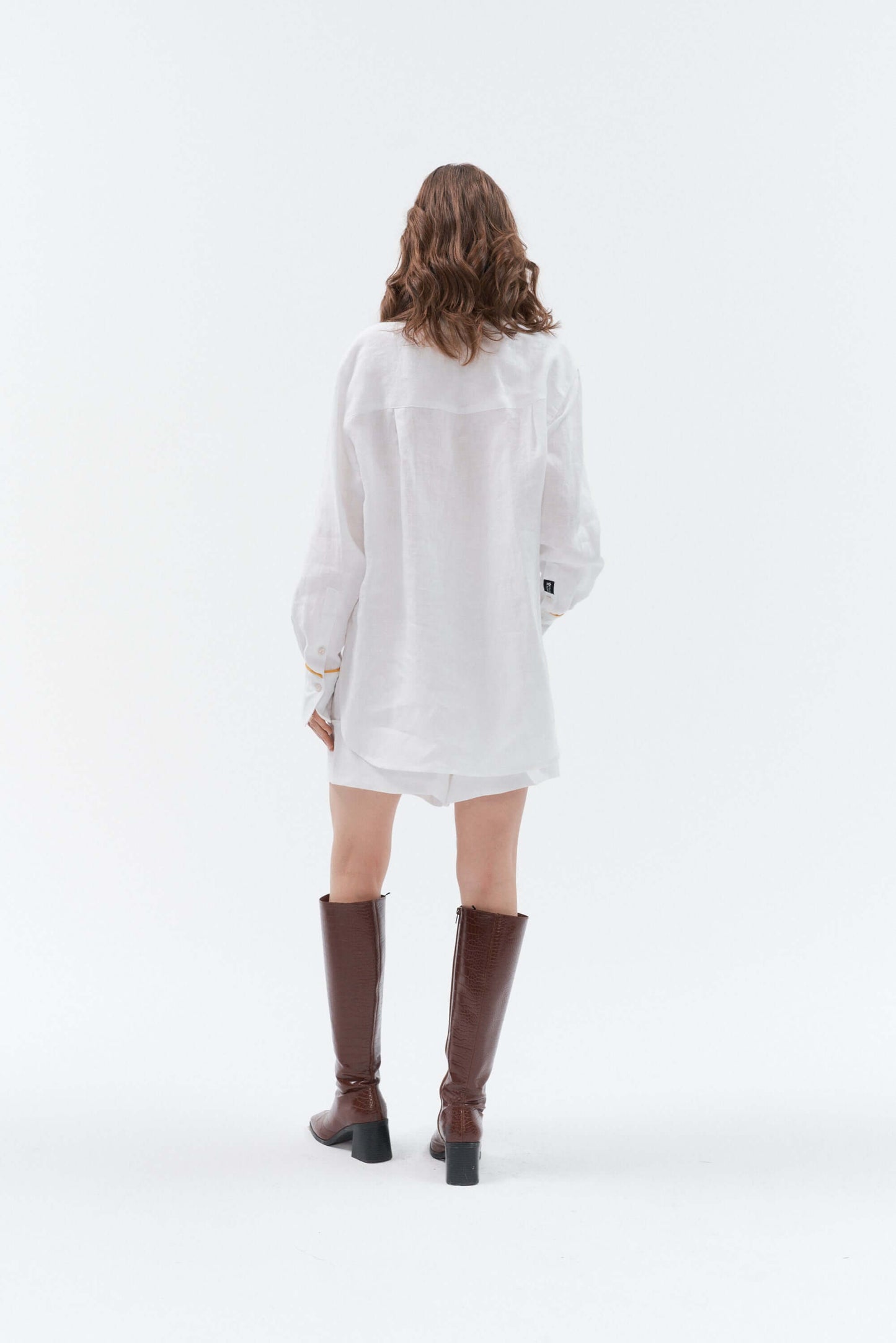 Back view of woman wearing white wide-leg linen shorts Bonita paired with the white Brillante long-sleeve oversized linen shirt with yellow accents, styled with brown high-heeled boots.