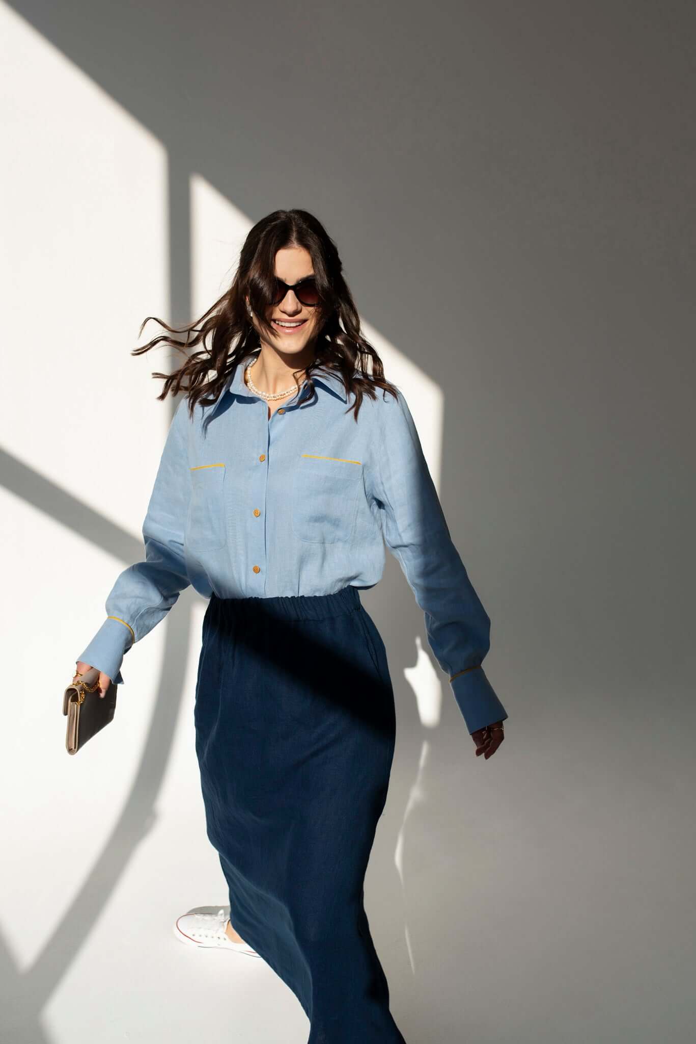 Woman wearing a light blue linen shirt and long navy blue linen skirt Soho against a white wall.