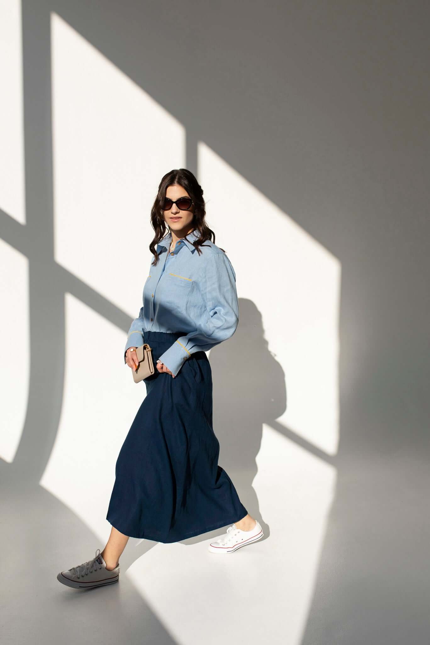 Woman wearing oversized linen shirt Brillante with yellow details and long navy skirt Soho standing in a sunlit room with shadows on the wall.