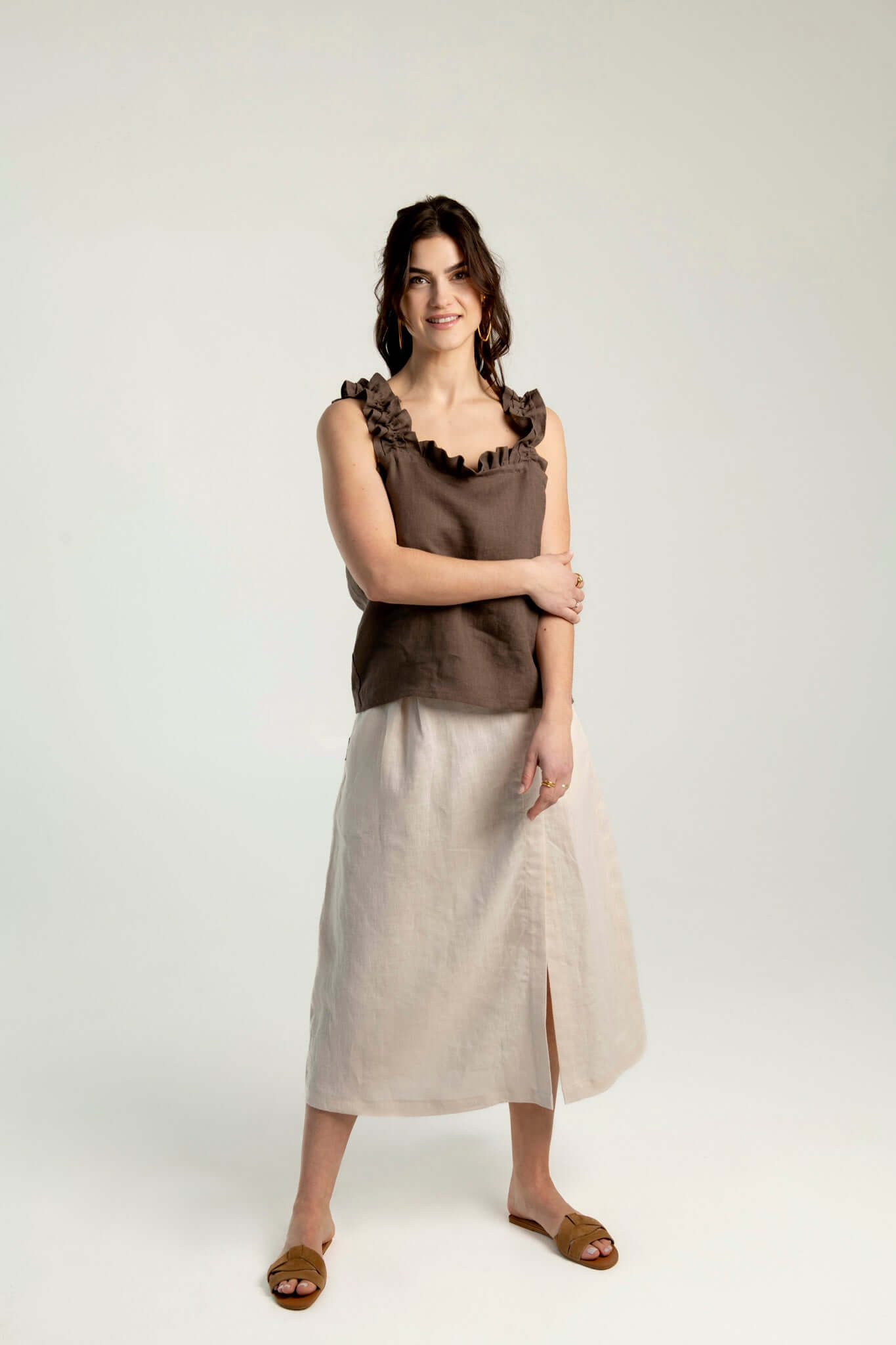 Front view of a woman wearing a brown linen top with ruffled straps paired with a beige midi linen skirt Madison with side slit – elegant, breathable summer outfit on a plain background.